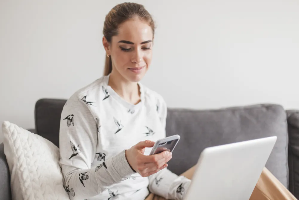 Frau sitzt mit Smartphone und Laptop auf dem Sofa